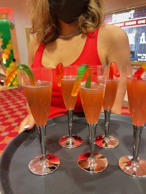 A woman wearing a mask is holding a tray of drinks with gummy worms in them.