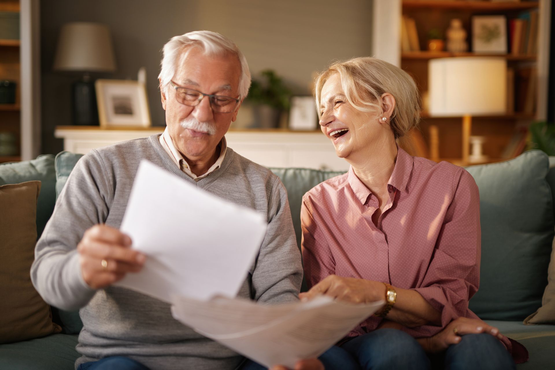 Elderly couple smiling and reviewing paperwork on a couch in a living room.