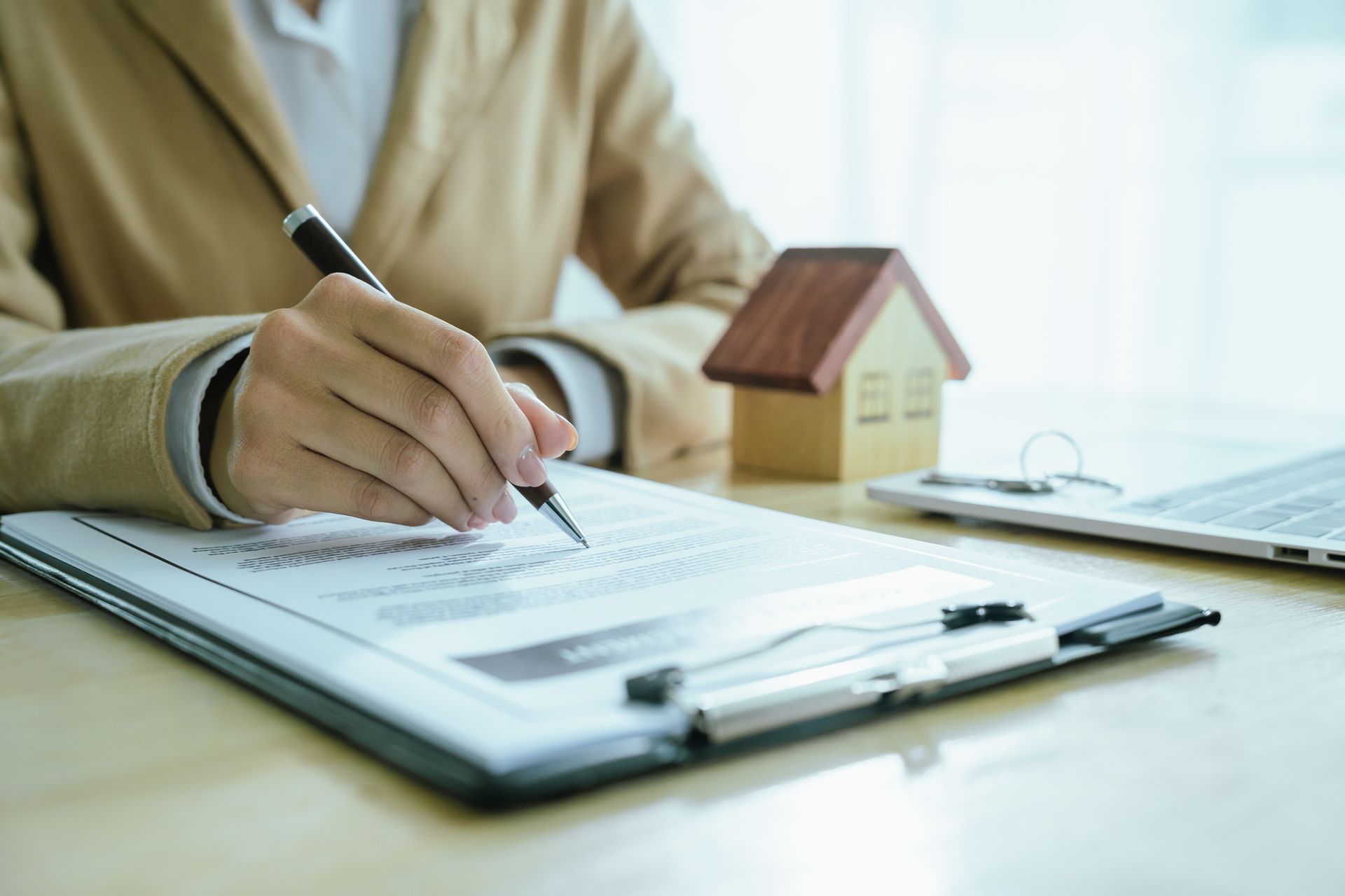 Person in tan jacket signing paperwork, small house model and laptop on desk.