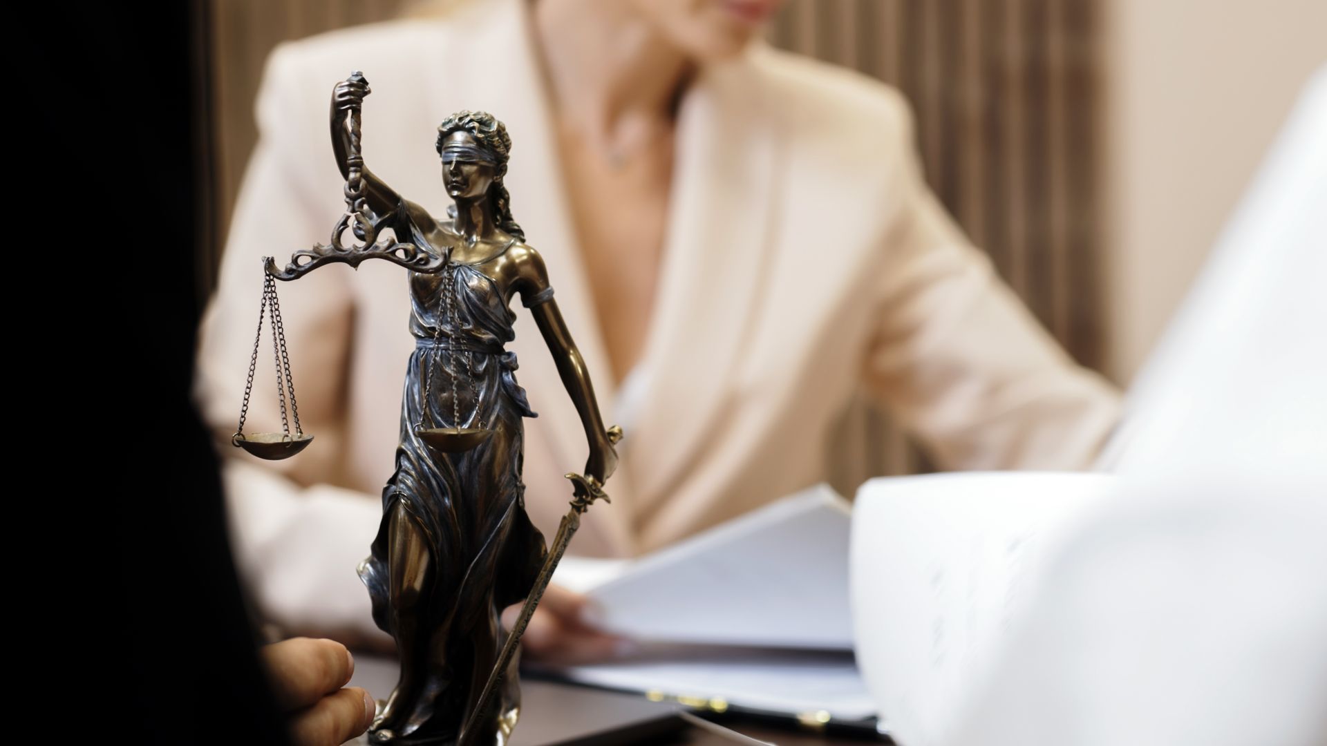 Statue of Lady Justice on a desk, woman in blazer in background, reviewing documents.