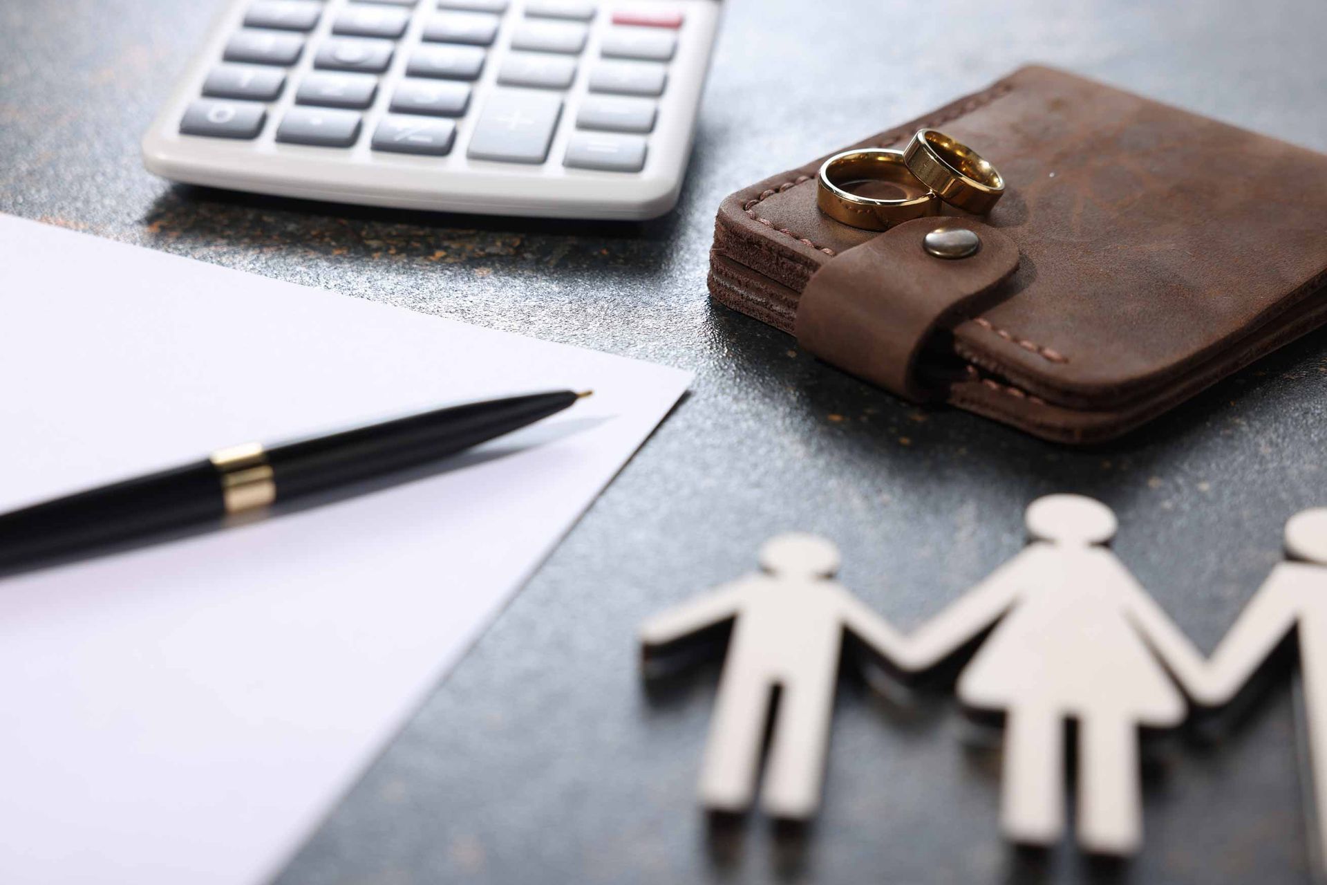 A calculator, a pen on a blank sheet of paper, a leather wallet with two gold wedding rings on top. A calculator, a pen on a blank sheet of paper, a leather wallet with two gold wedding rings on top.