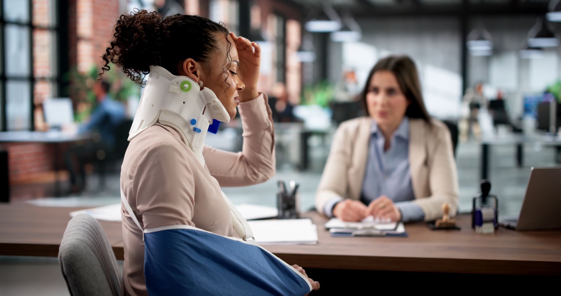 Woman with neck brace and arm sling at desk with another person in office.