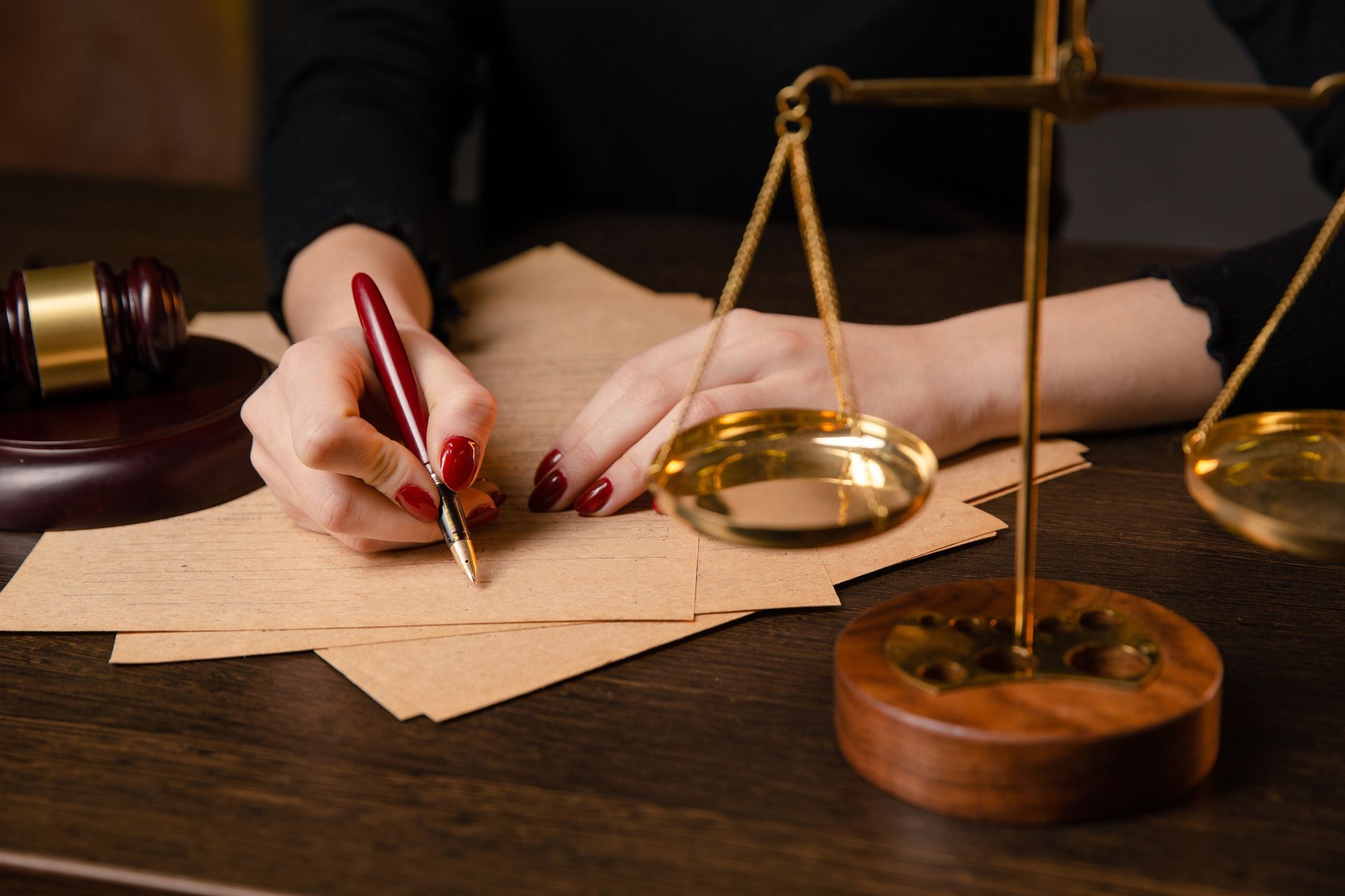 A person writing with a pen next to a justice scale and a gavel on a wooden desk.