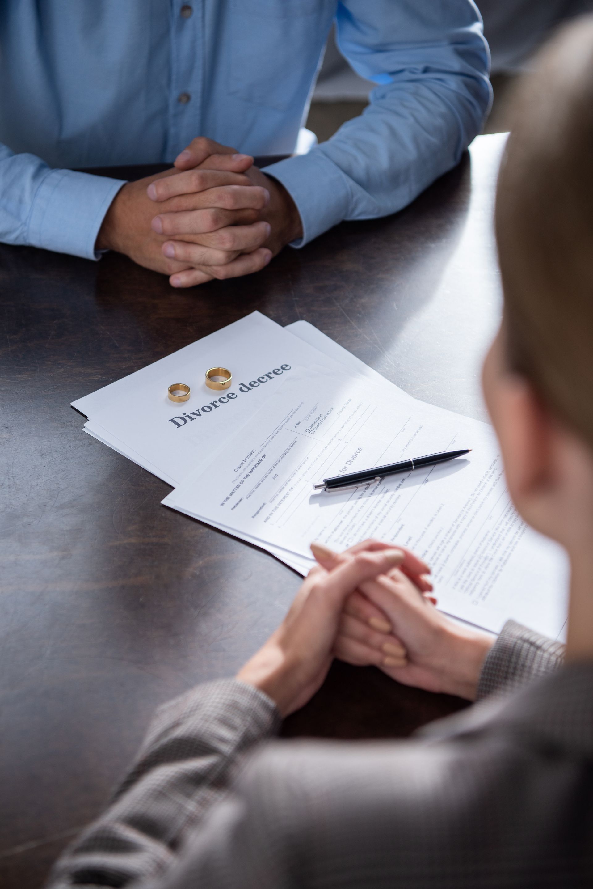 Divorce papers with rings, a pen, and two people at a table, hands clasped. Divorce papers with rings, a pen, and two people at a table, hands clasped.