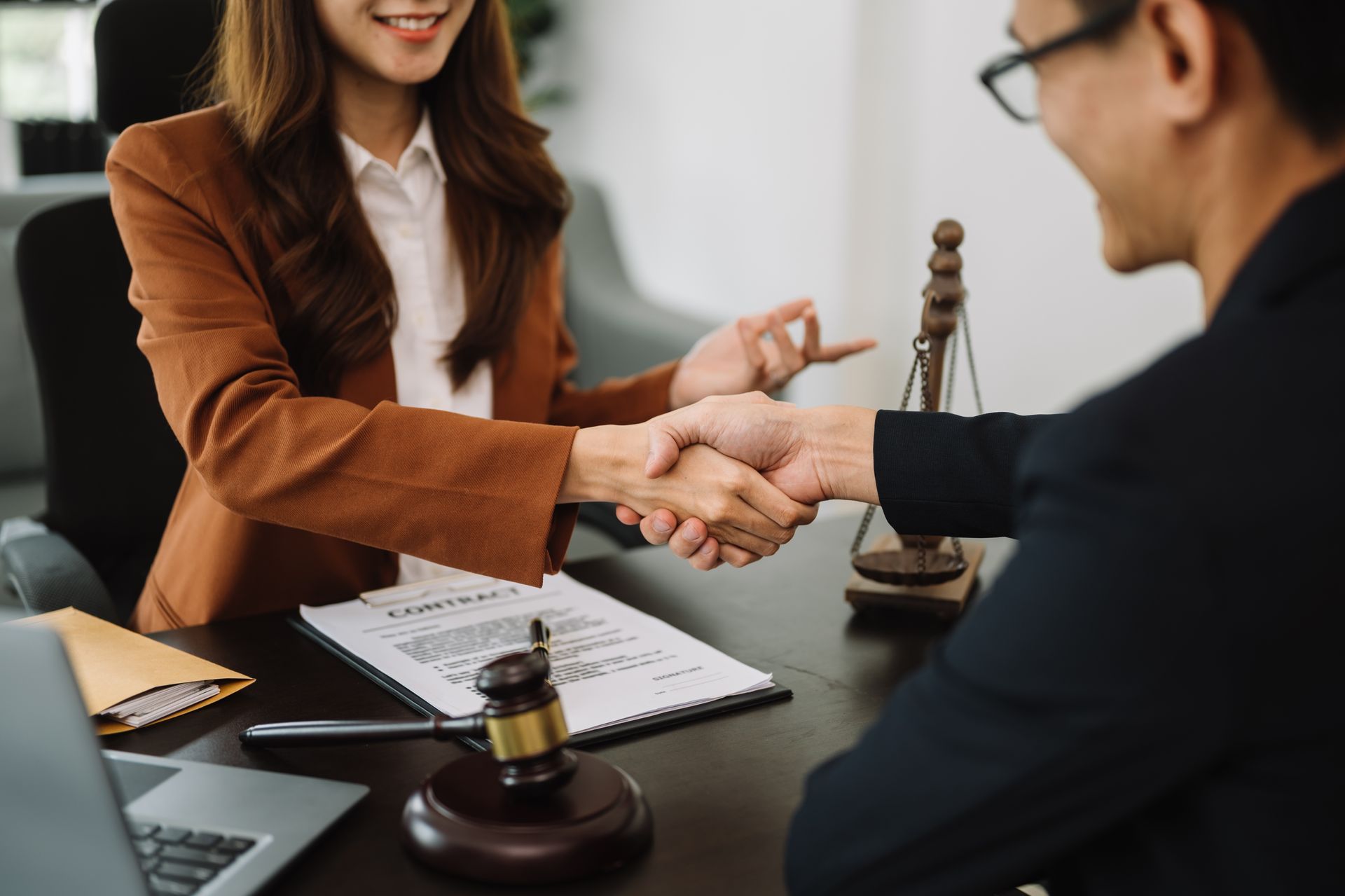 A woman in a brown blazer shakes hands with a man, formal office setting.