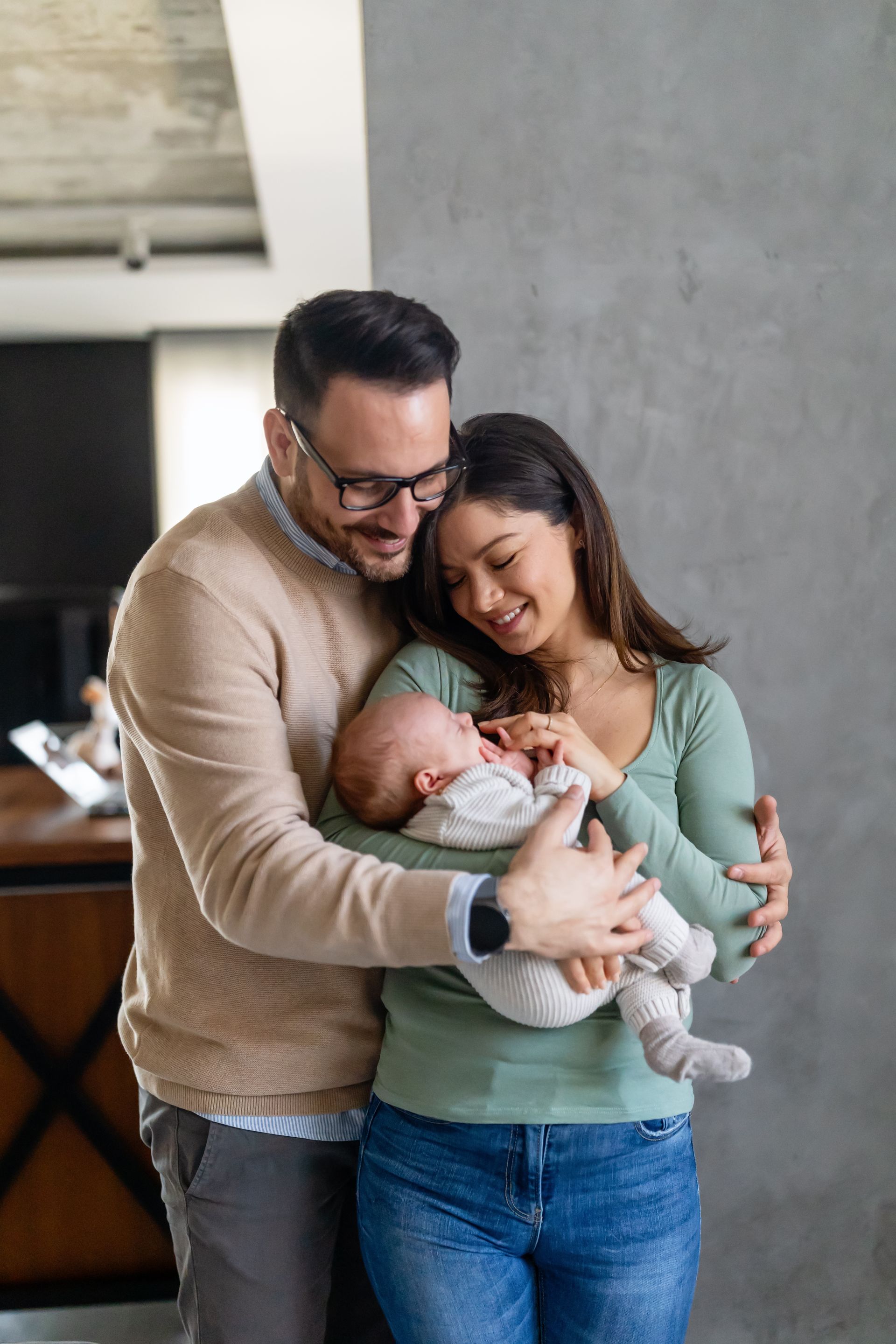 Couple smiling, holding baby. Man in glasses, tan sweater. Woman in green shirt, jeans. Gray wall background. Couple smiling, holding baby. Man in glasses, tan sweater. Woman in green shirt, jeans. Gray wall background.