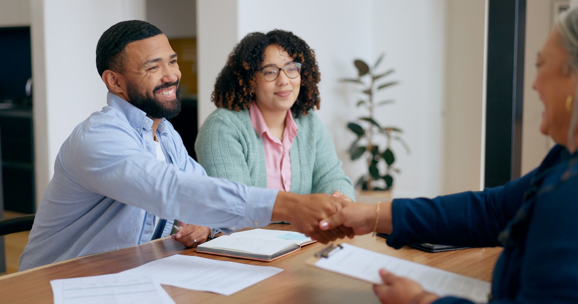 Man shaking hands with another person at a table, woman watches. Papers on table.