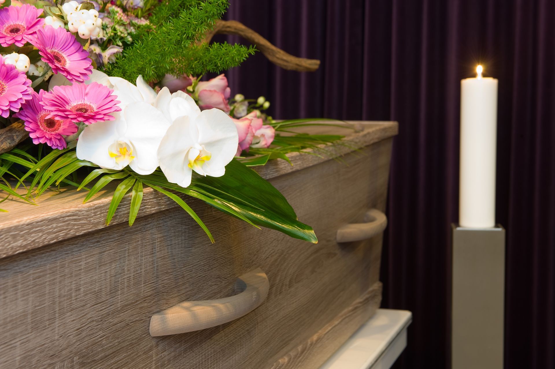 Coffin with flowers and a lit candle in front of a purple backdrop.