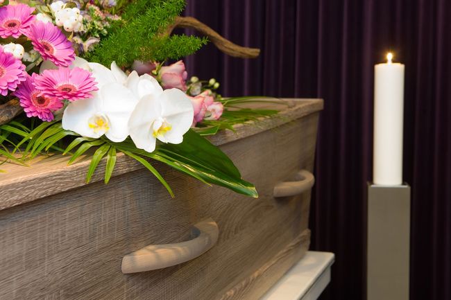 Coffin with flowers and a lit candle in front of a purple backdrop.