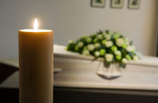 Candle burning brightly in front of a closed white coffin with floral arrangement, soft lighting.