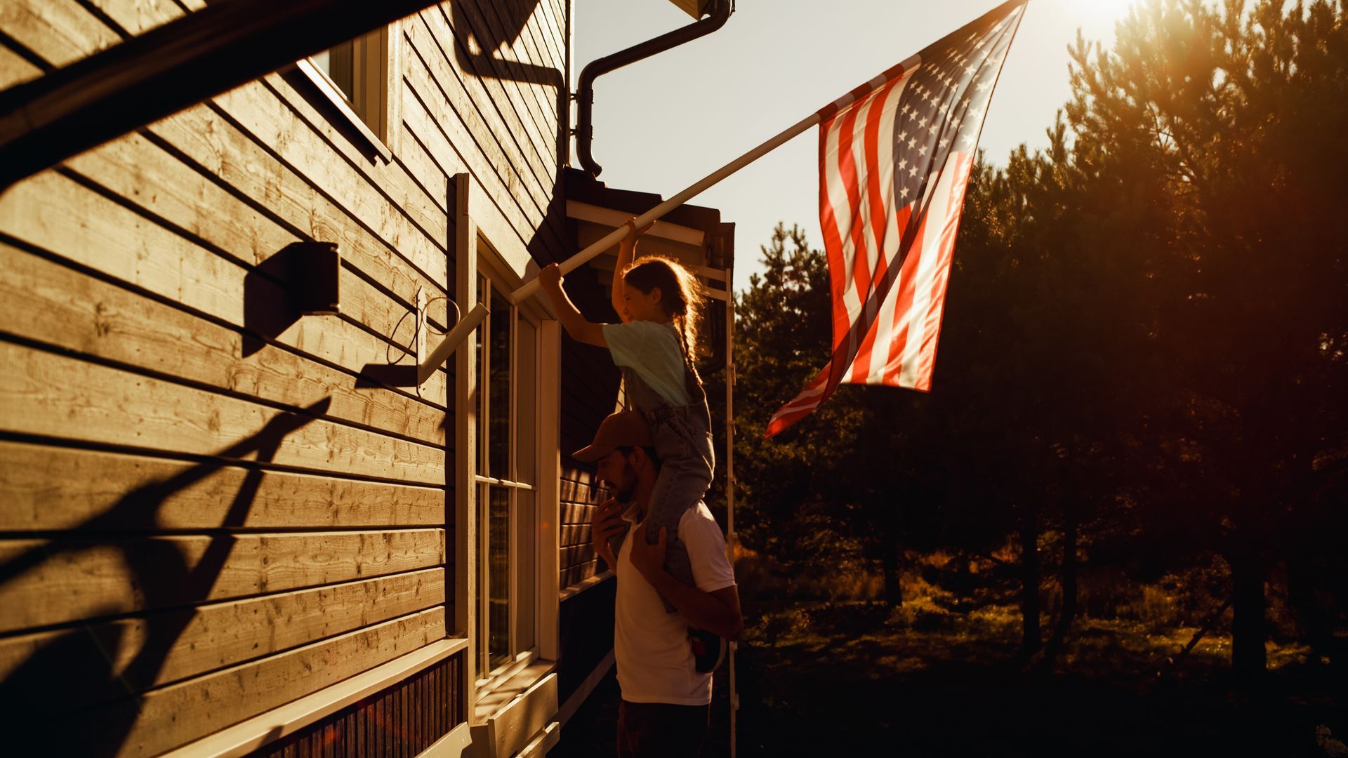 Two people on a porch, one reaching toward an American flag at sunset.