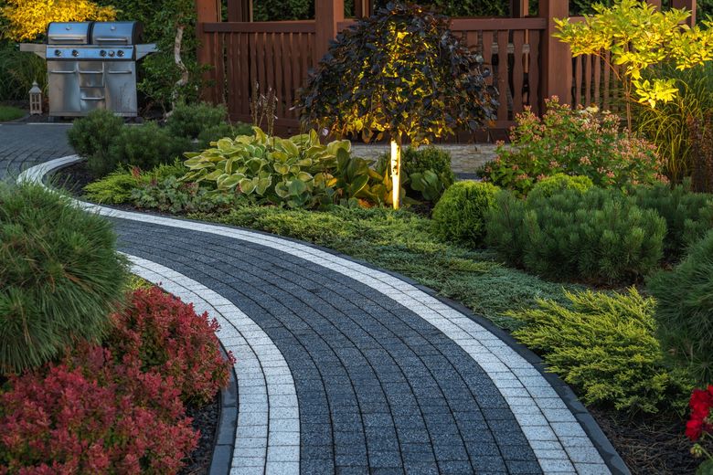 Curving brick path through a garden with shrubs, leading to a grill and gazebo, lit by warm lights.