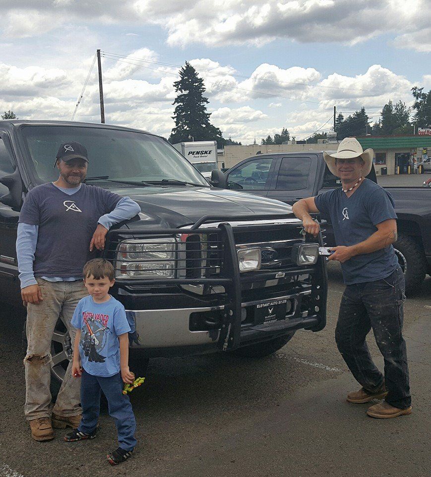 Three people and a child pose by a black truck with a guard; sunny, outdoor setting.
