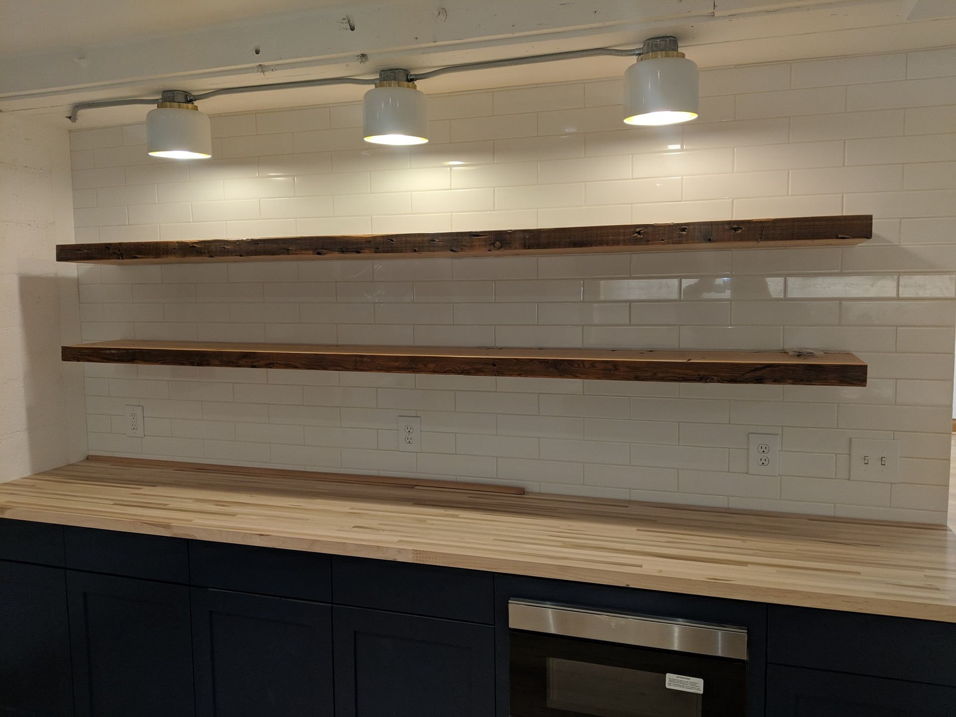 Two rustic wooden floating shelves mounted on a white subway tile wall above a dark kitchen cabinet and wood countertop.