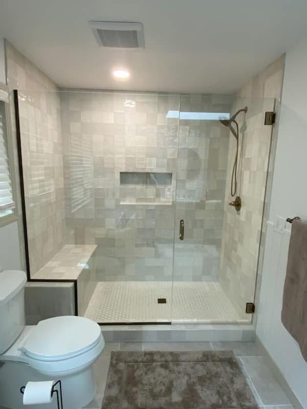 A modern walk-in shower with light tile walls, a glass door, a built-in bench, and a gold shower head fixture.