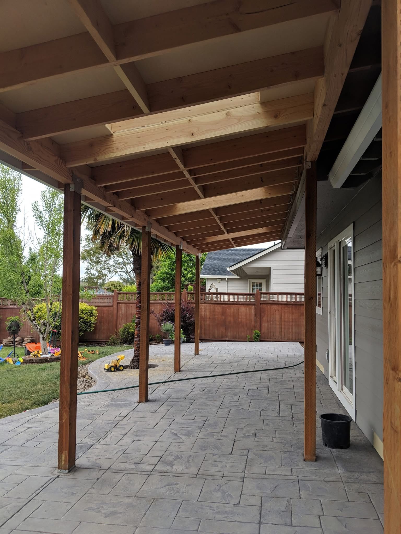 A wooden patio cover extends from a gray house over a gray stamped concrete patio in a backyard with a wooden fence.