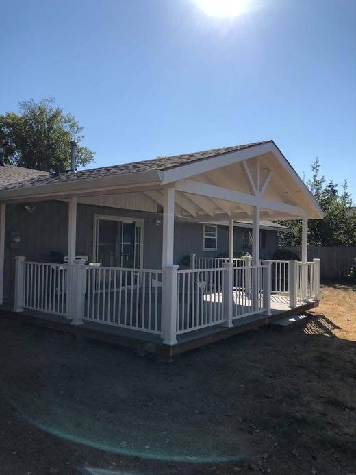 A grey house with a newly constructed, white-railed covered wooden deck and a gabled roof against a bright blue sky.