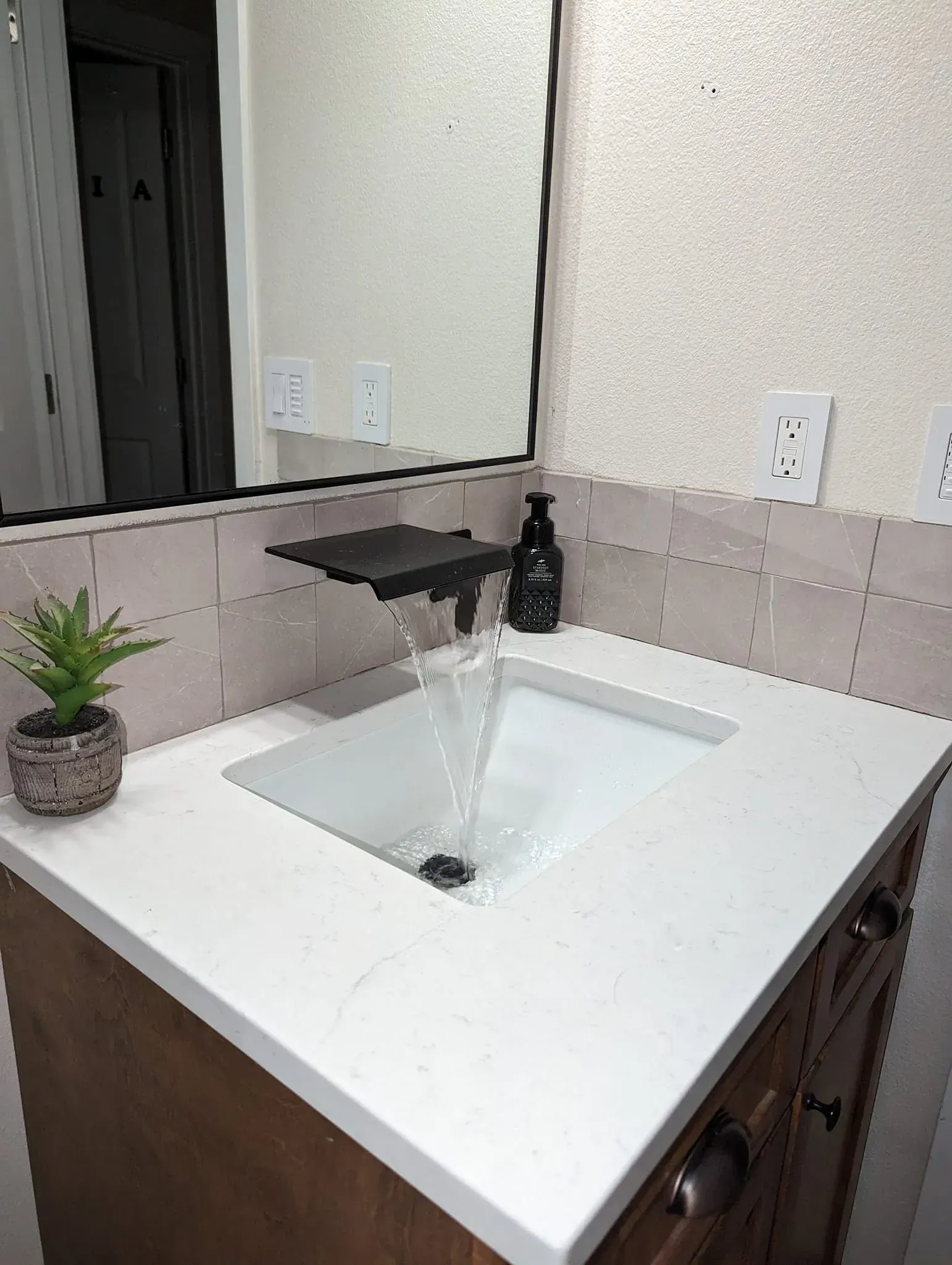 Bathroom vanity with running water, white countertop, black faucet, mirror, soap dispenser, and small plant.