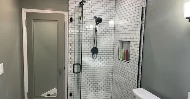 Bathroom with white tiled shower, black fixtures, glass door, and gray walls.