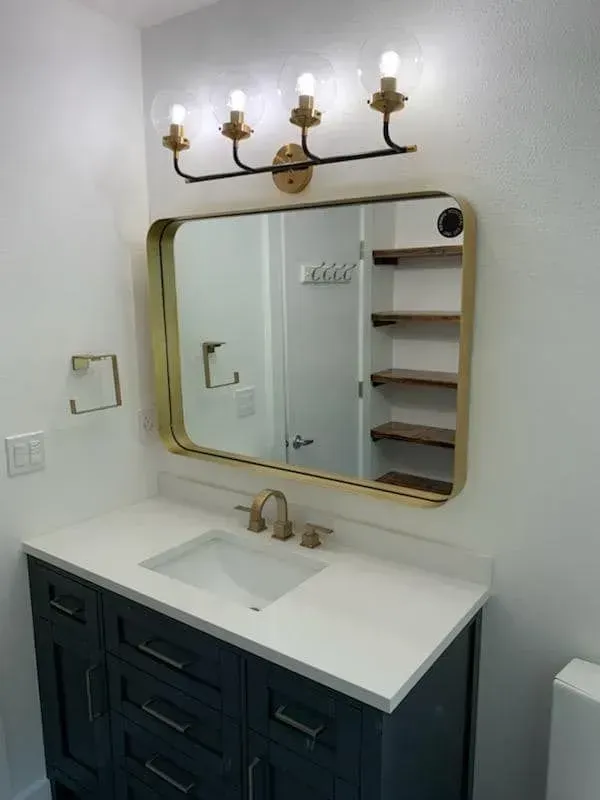 Bathroom with navy vanity, gold-framed mirror, and brass fixtures. White walls, wood shelves visible in the mirror.