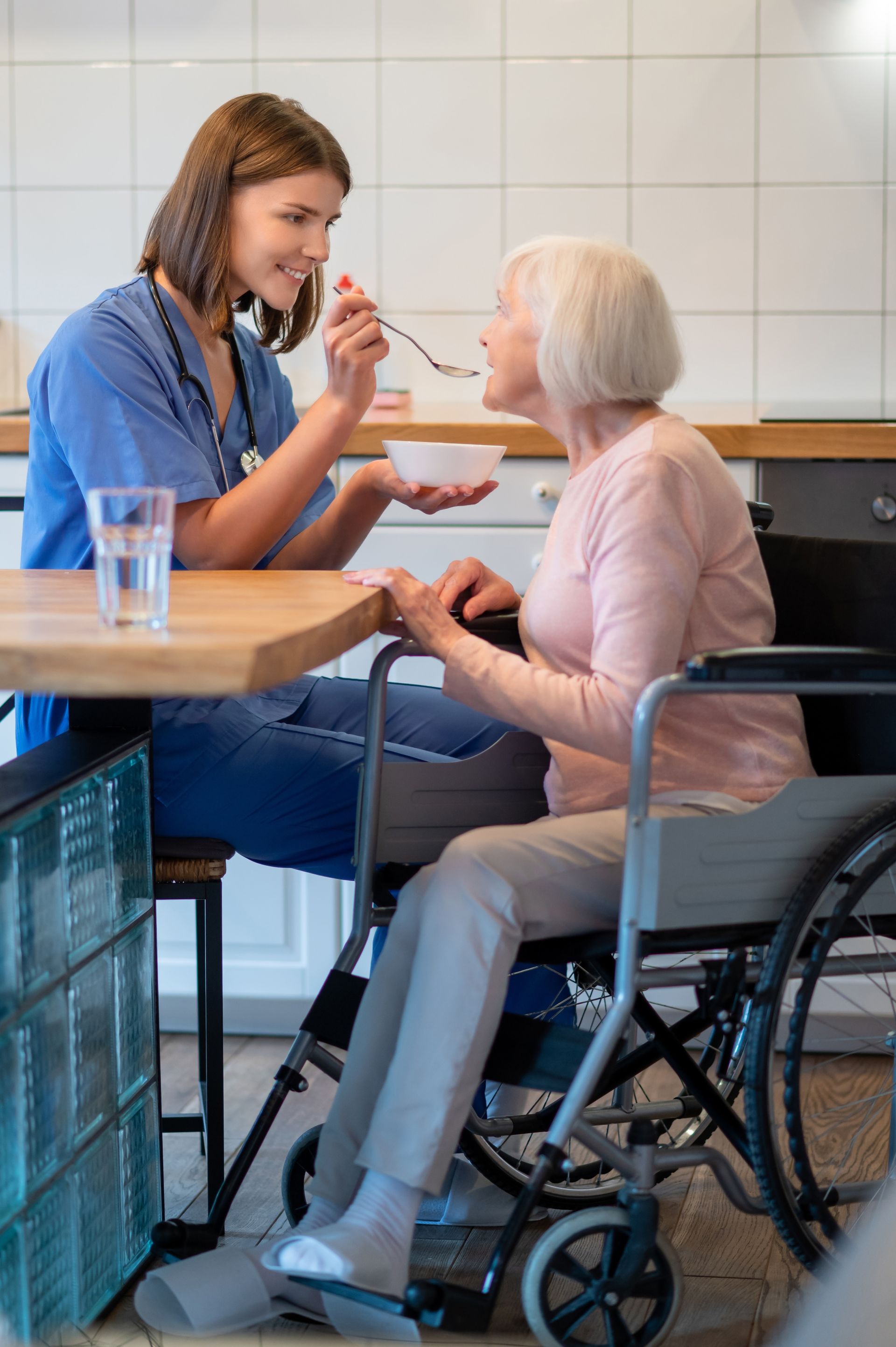 A caregiver in blue scrubs feeds a person sitting in a wheelchair at a kitchen table.