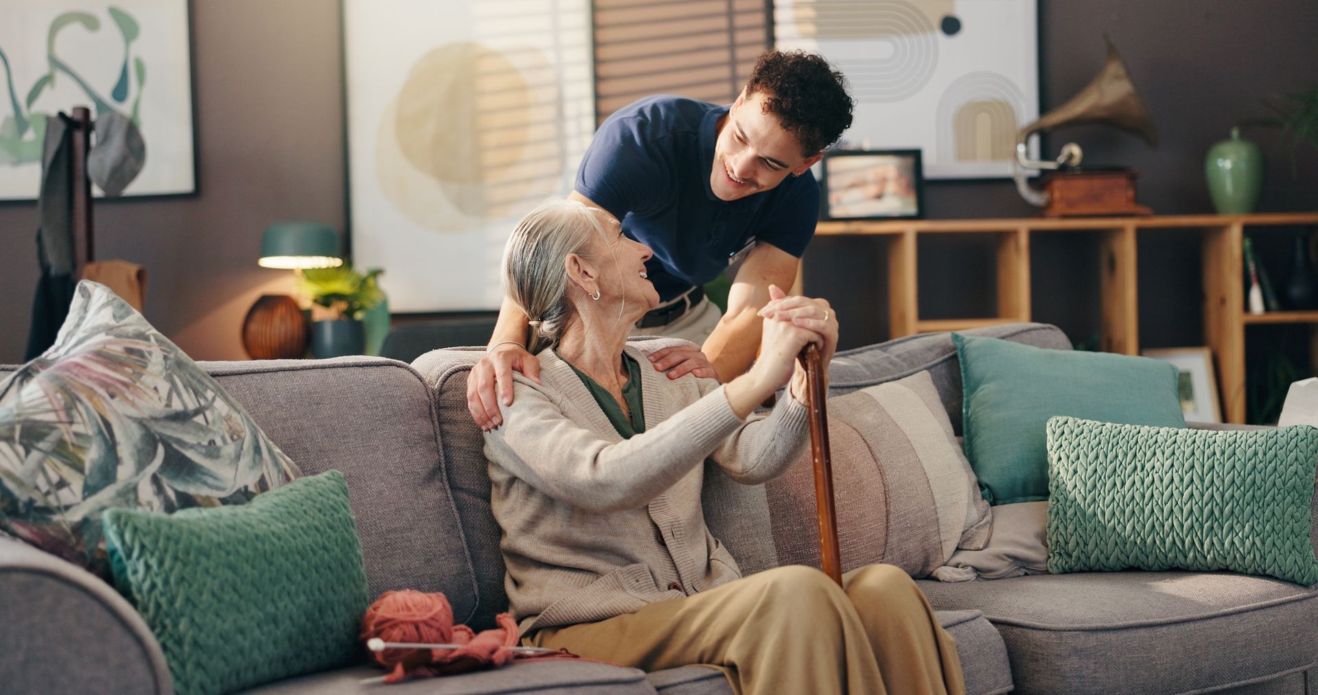 A person leans over a sofa, smiling kindly at an individual sitting on the couch while holding a wooden cane.