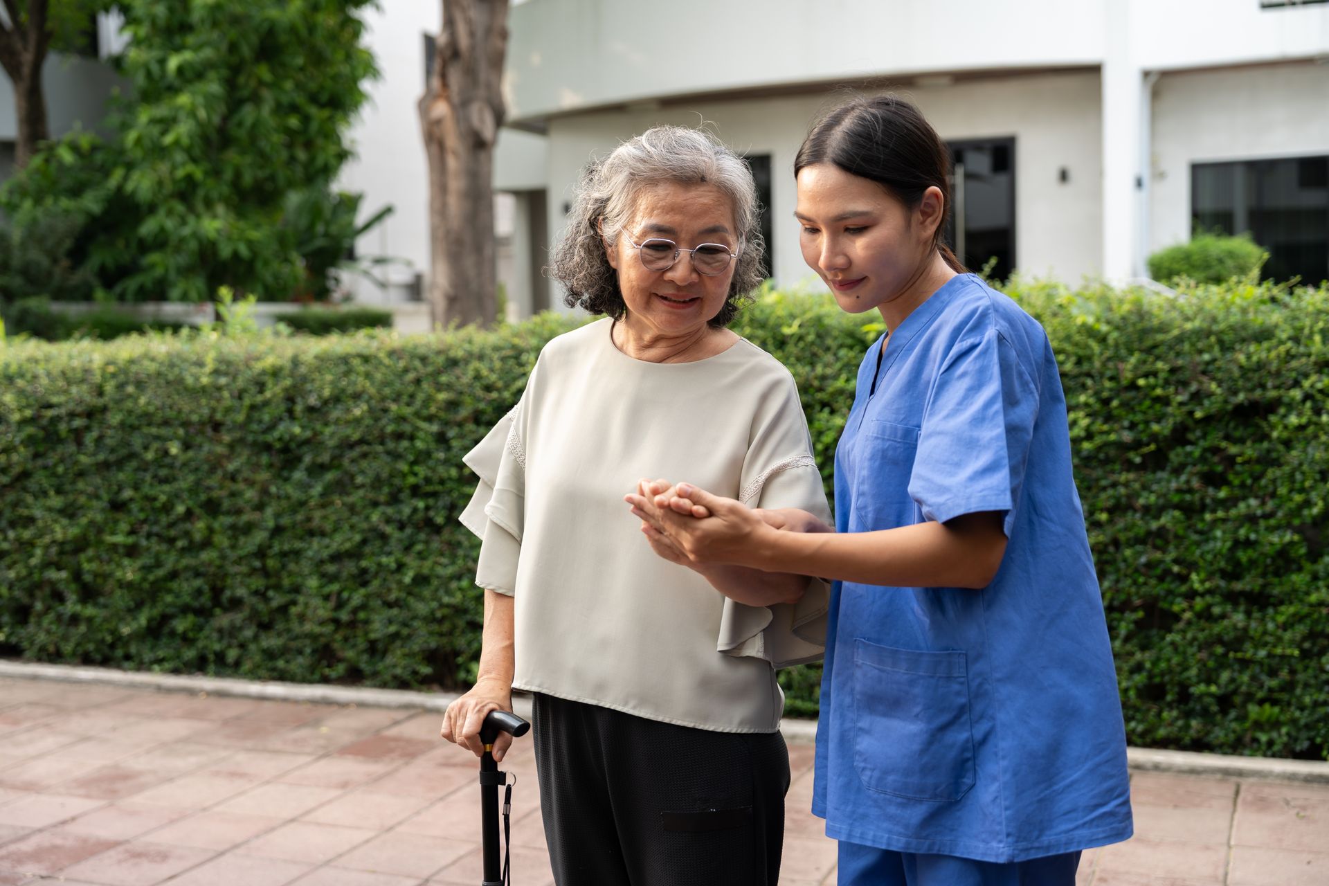 A caregiver in blue scrubs helps a person with a cane look at a phone outdoors.