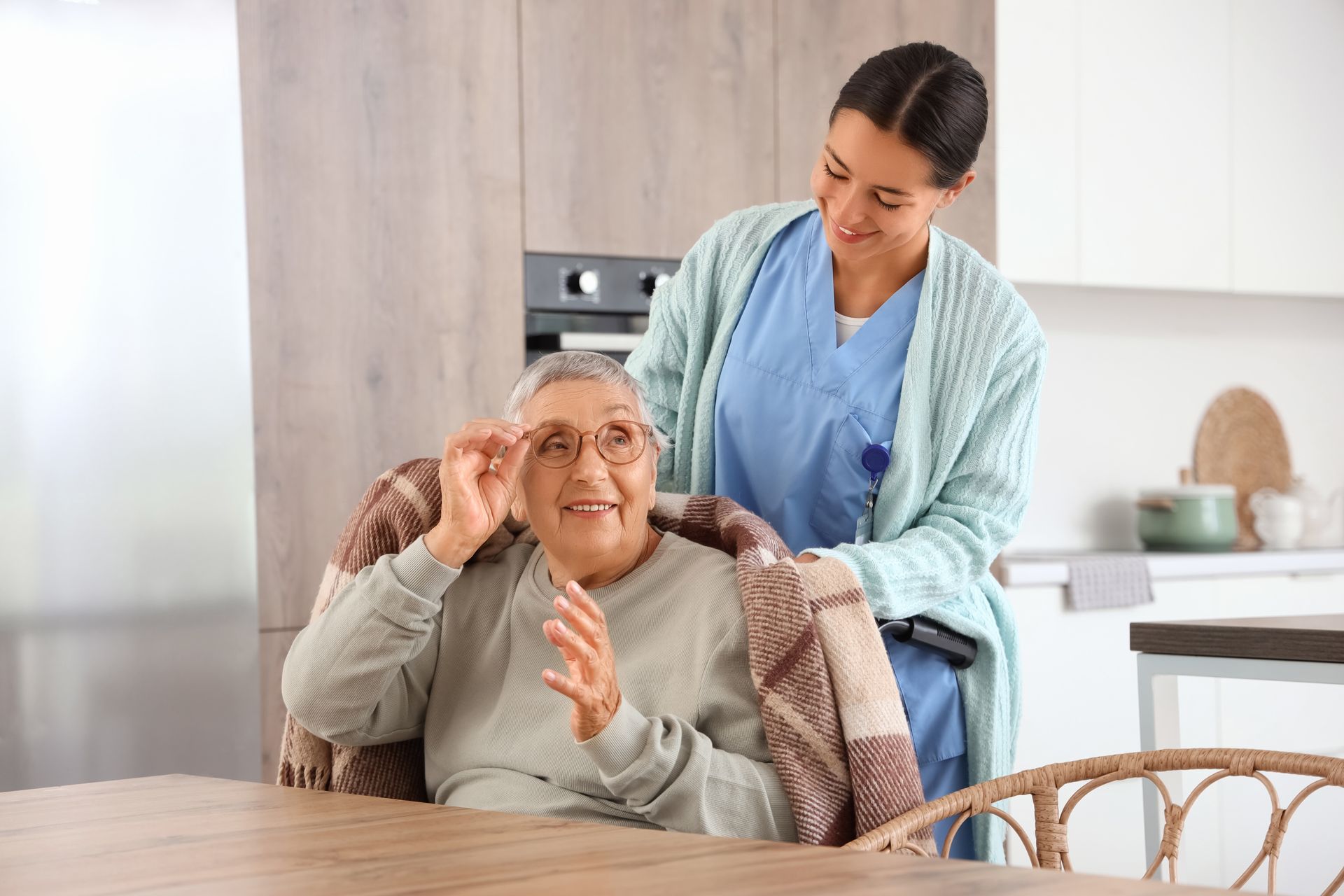 A caregiver smiles while placing a blanket over an elderly person, who is smiling and adjusting their glasses.