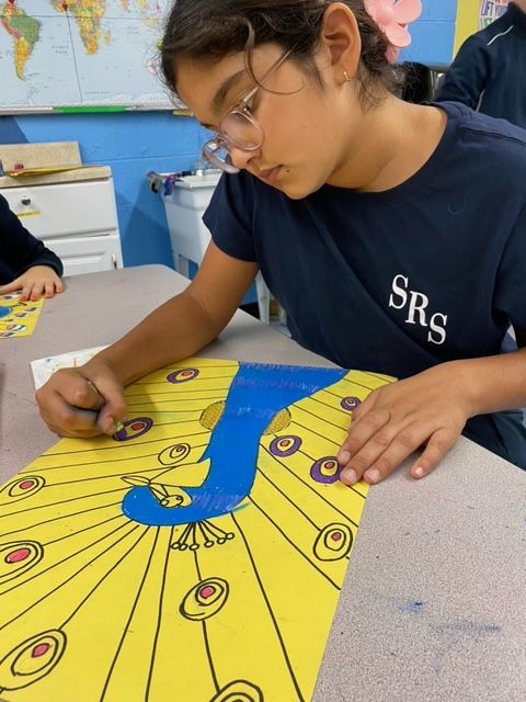 A middle school student in a navy T-shirt that has the school initials SRS on it colors in a peacock she in drawing in art class.