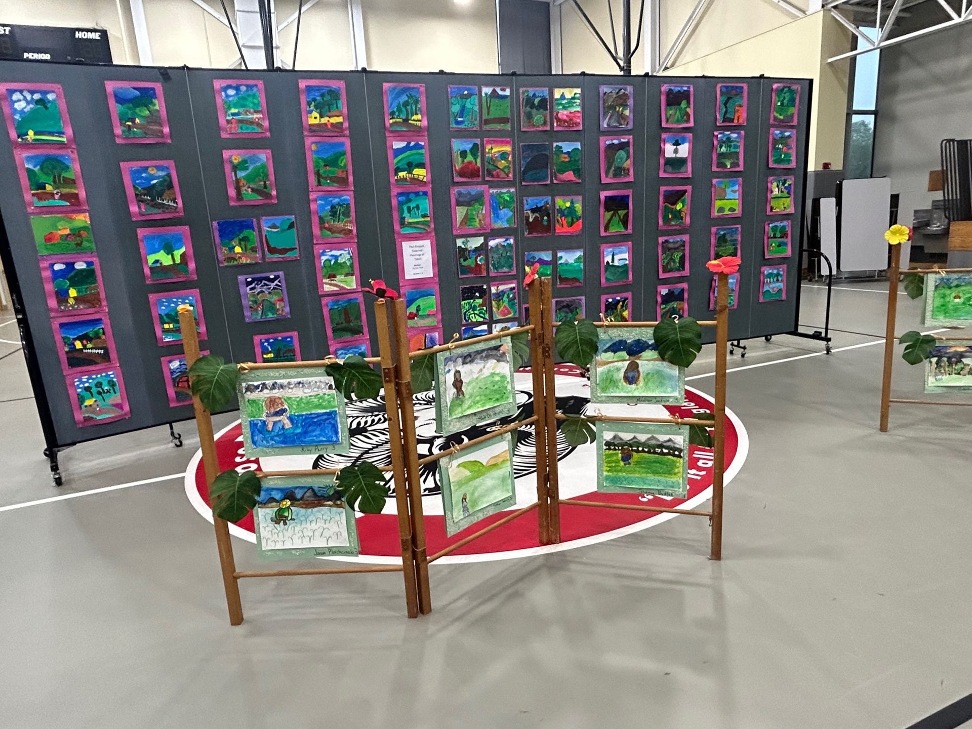 Photo shows a wall divider with student paintings of all the same style mounted on it. In the foreground a smaller divider displays several more paintings