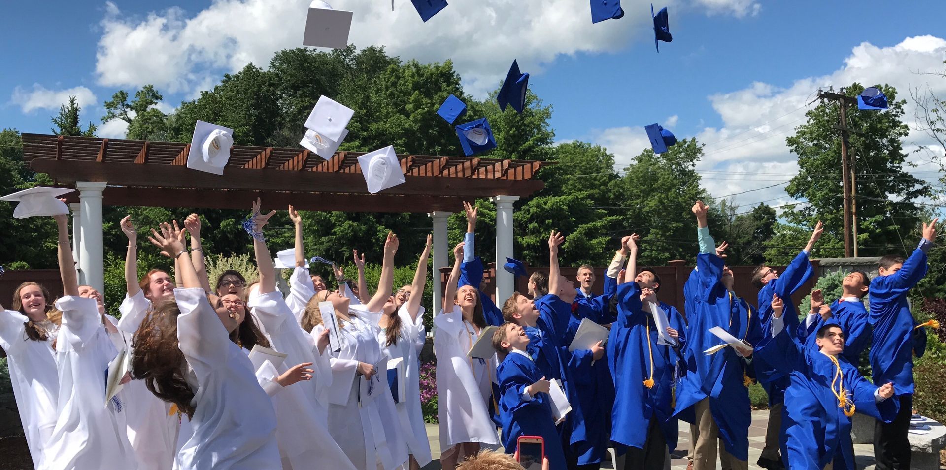 Girls in white gowns and boys in blue gowns toss their graduation mortar board caps in the air and cheer.