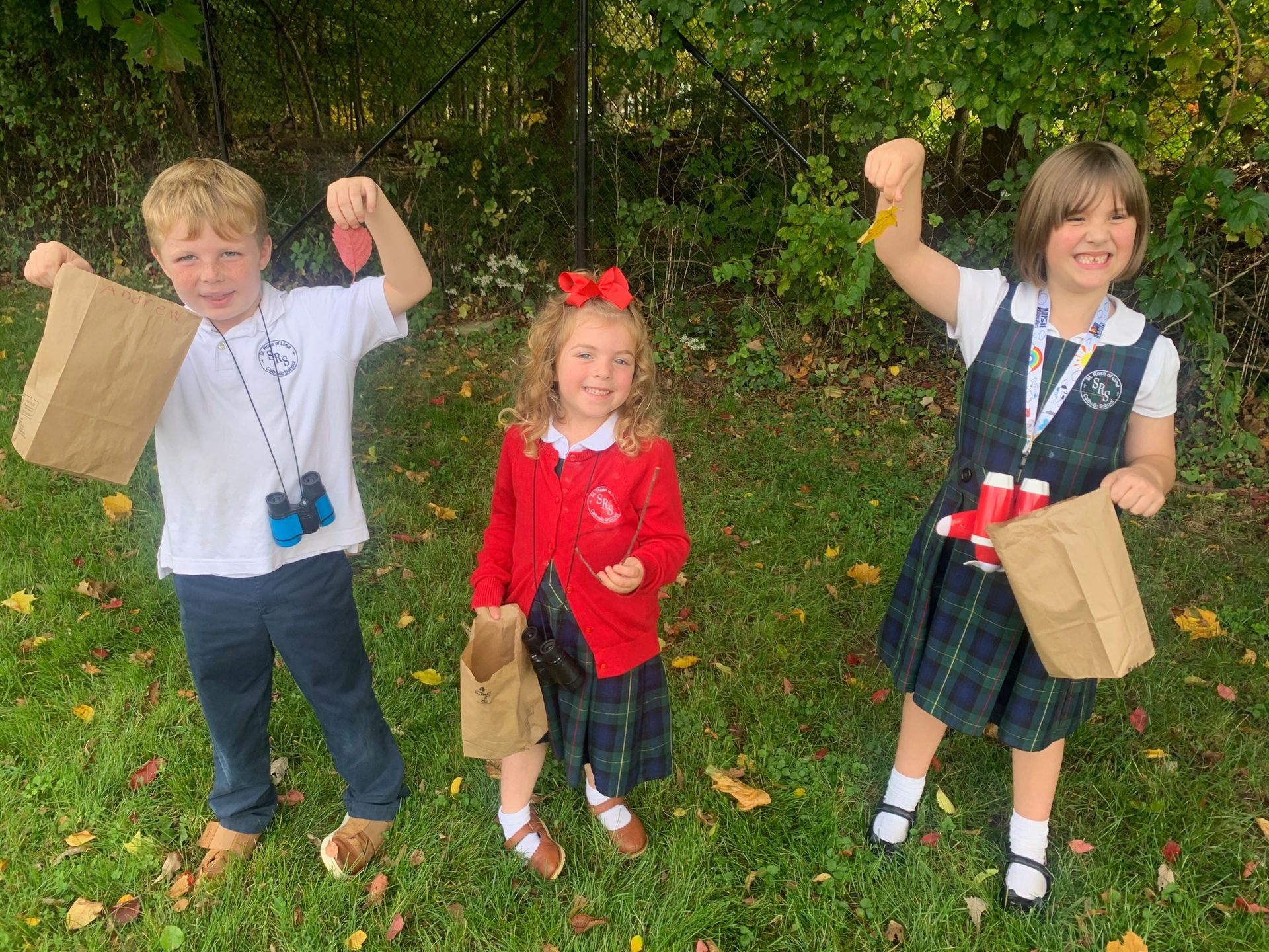 Three elementary students in their school uniforms hold up fall leaves they are collecting outside.