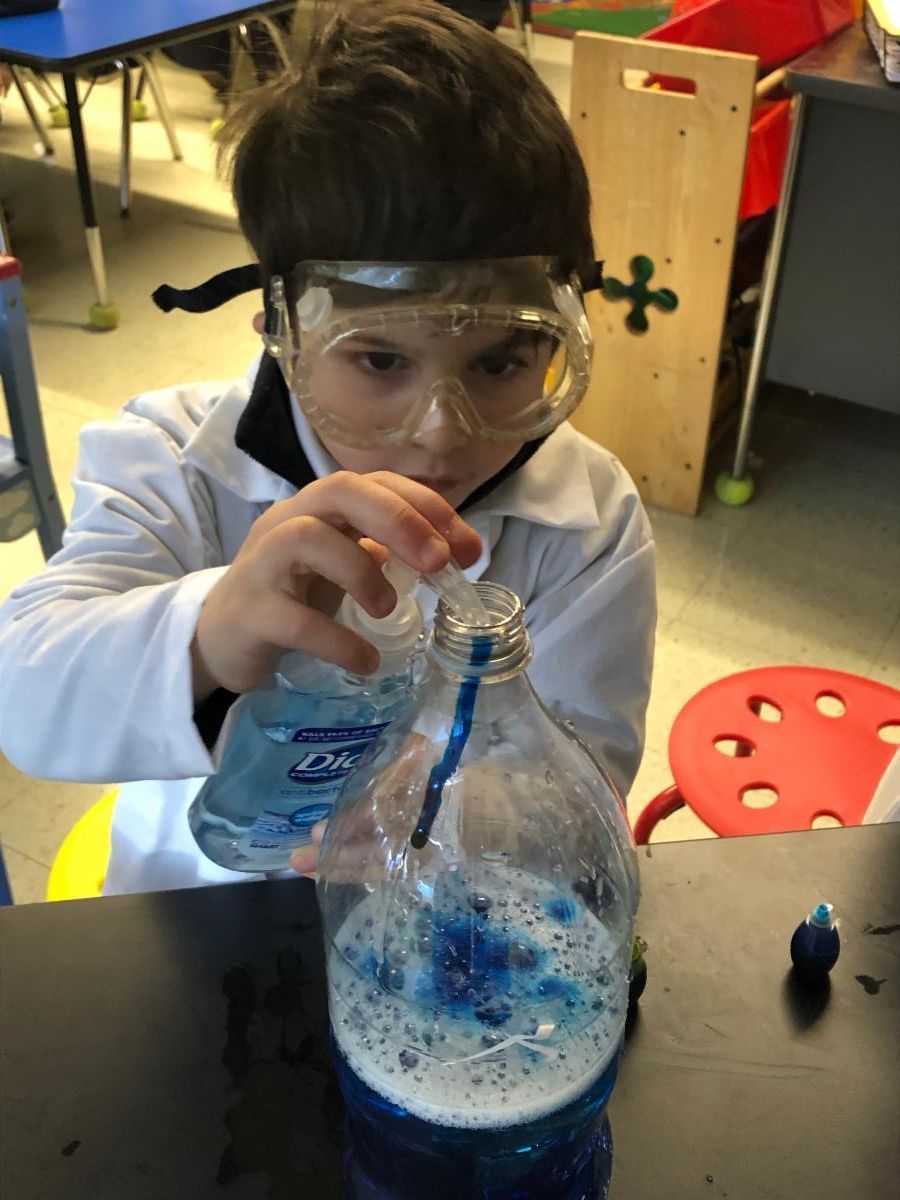 A Kindergarten student wears safety goggles as he adds soap to an Erlenmeyer flask that has a blue liquid inside.