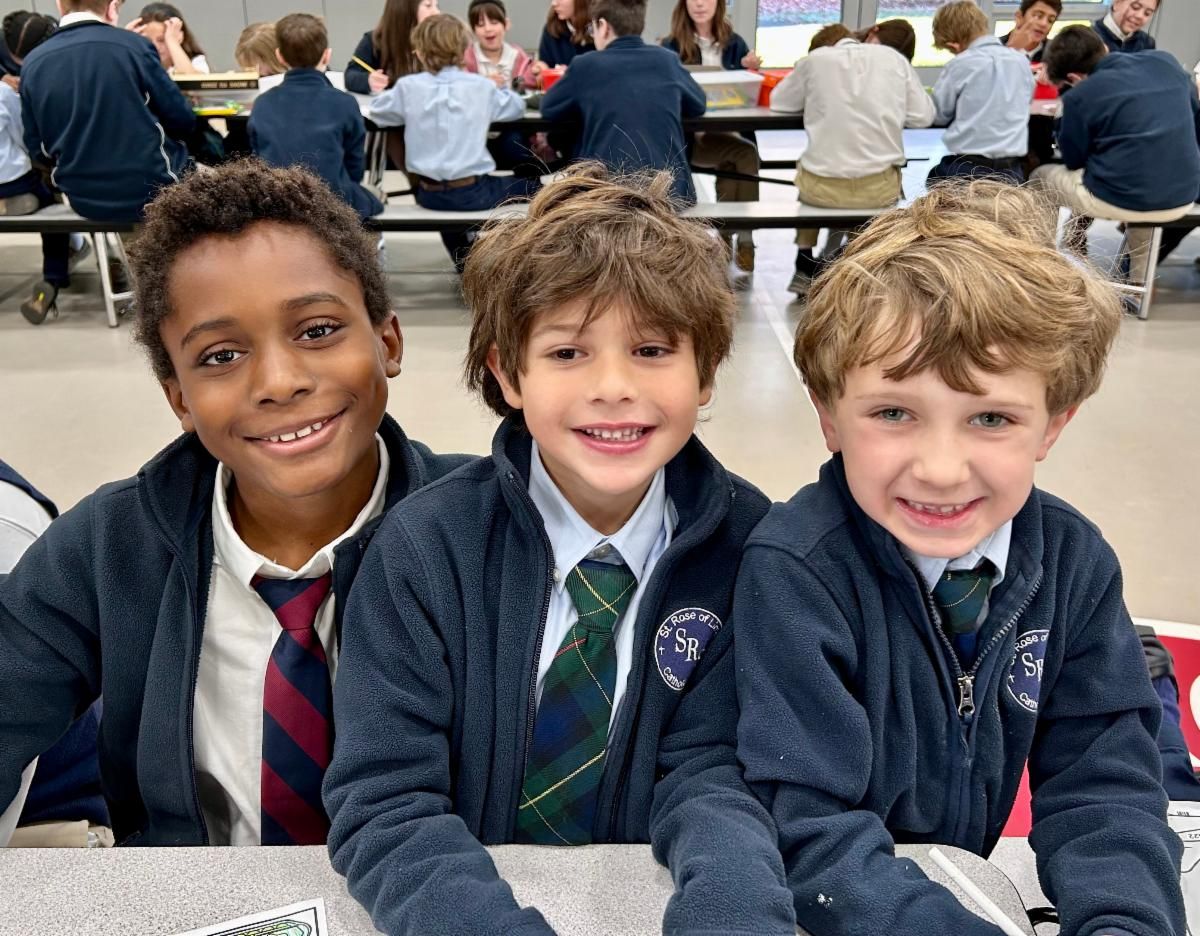 Three boys in St. Rose School uniforms sit at a cafeteria table and smile at the camera.