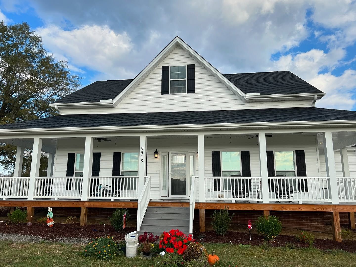 White farmhouse with black shutters and a wraparound porch under a cloudy sky.
