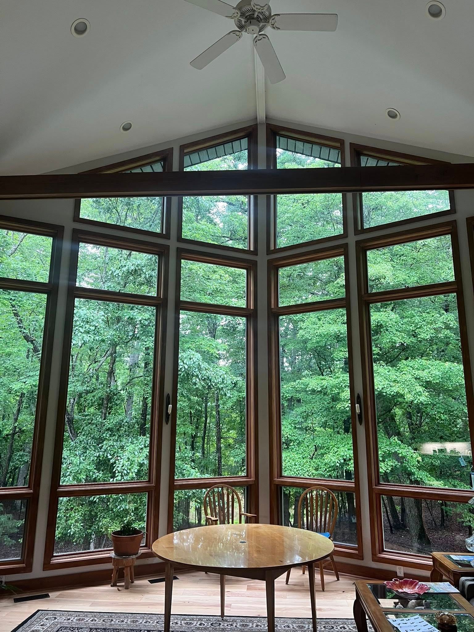 Interior view of a room with large windows overlooking a lush green forest, ceiling fan, and wooden furniture.