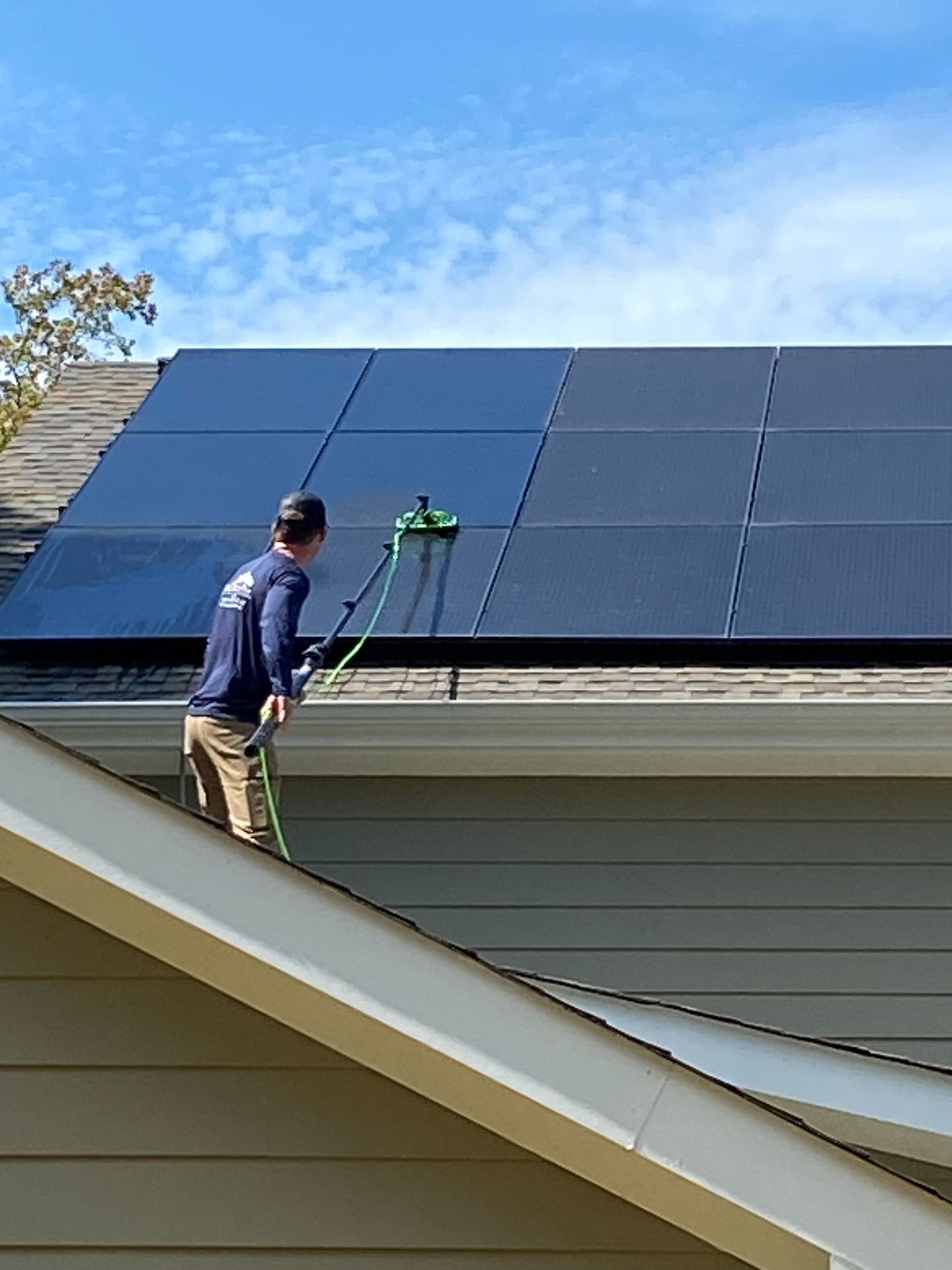 Person cleaning solar panels on a roof with a green brush and water hose; blue sky.