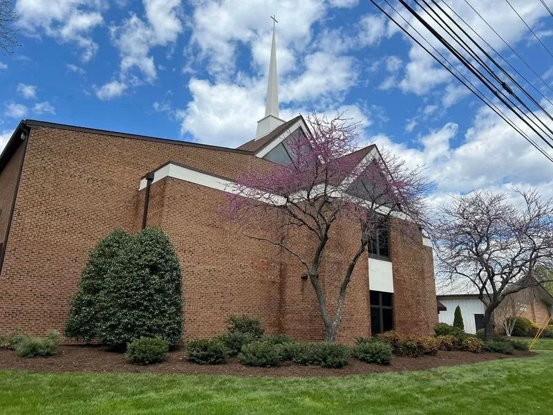 Brick church building with a steeple and a tree with pink blossoms under a blue sky.