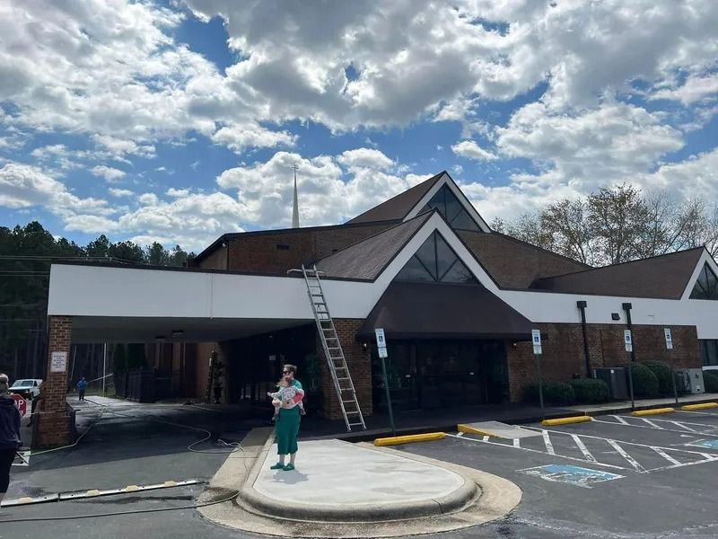 Exterior view of a church with a ladder leaning on the roof and a person standing on a concrete island. Cloudy sky.