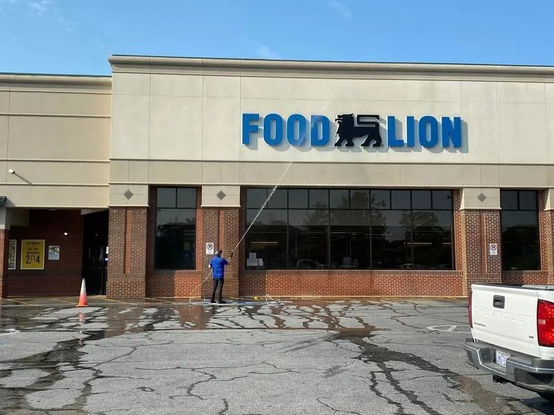 Person power washing the exterior of a Food Lion supermarket. Blue signage on a beige building, wet pavement.