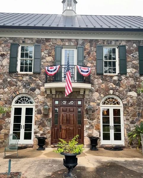 Stone building with American flag, balcony, and ornate door. Green shutters and a small cupola.