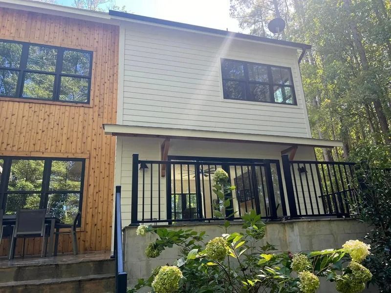 Two-story house with light siding and wood paneling, black framed windows, and a black railing deck, surrounded by trees.