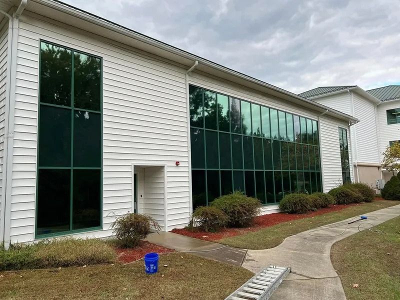 White building with large tinted windows. Brown bushes and sidewalk in front. Cloudy sky.