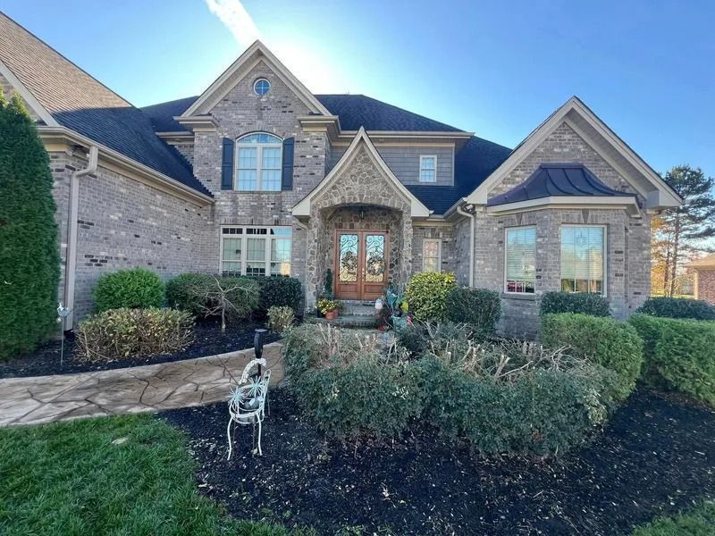 Brick home with dark roof, arched entry, and manicured landscaping under a blue sky.