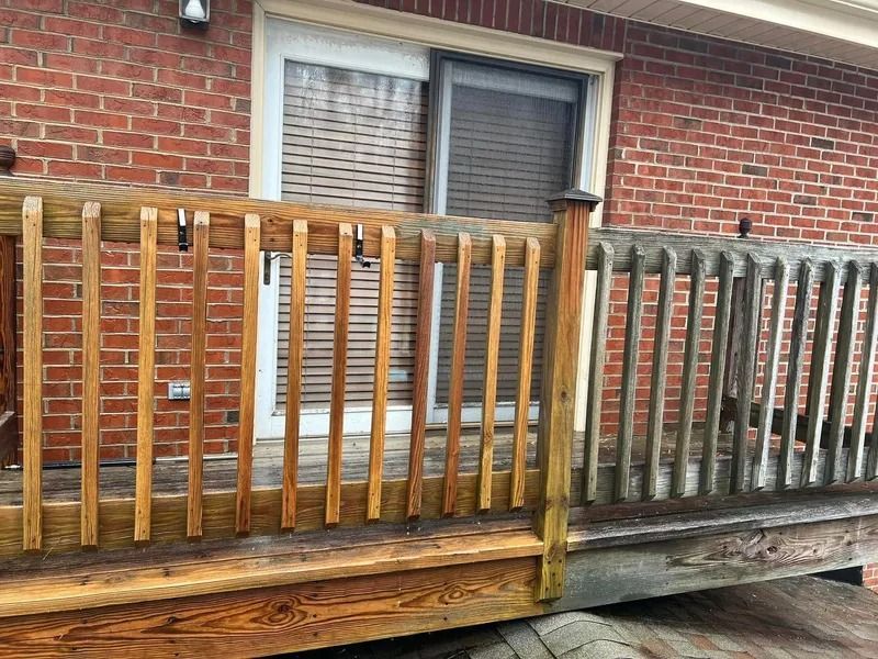 Wooden deck railing partially cleaned, showing before and after power washing. Red brick building in background.