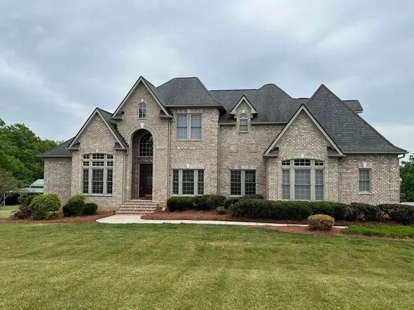 Large brick house with dark roof, windows, and manicured lawn under a cloudy sky.