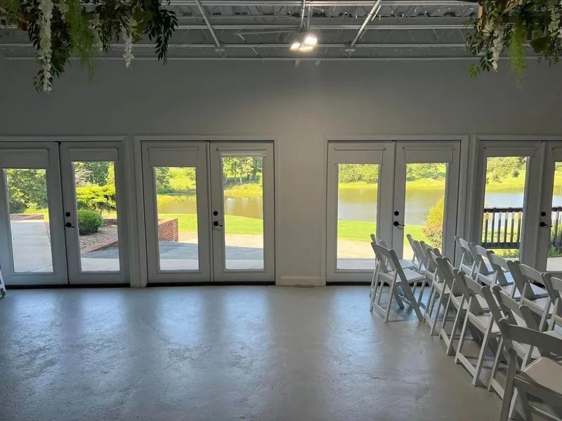 Bright, empty event space with white doors overlooking a lake; white folding chairs are set up.