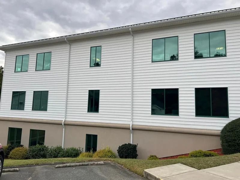 Two-story white building with green-tinted windows, tan base, and green bushes in front. Overcast sky.