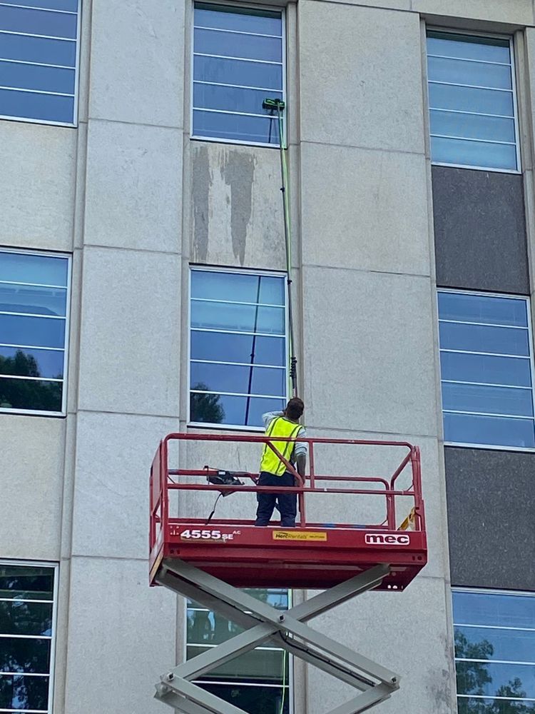 Person on a red lift washes windows on a multi-story building using a long-handled brush.