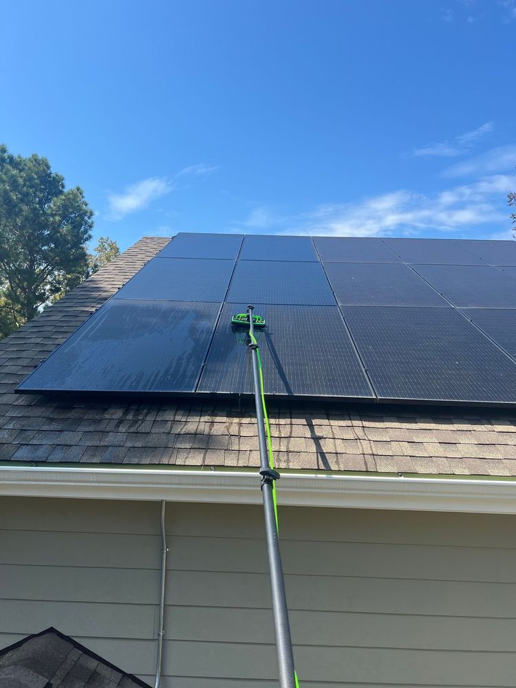 A person cleans solar panels on a roof with a long-handled brush under a blue sky.